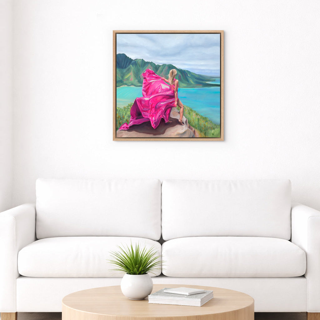 Framed artwork of a woman in a pink dress standing on a mountain in Hawaii, overlooking the ocean, on a white wall above a modern white sofa with a wooden coffee table.