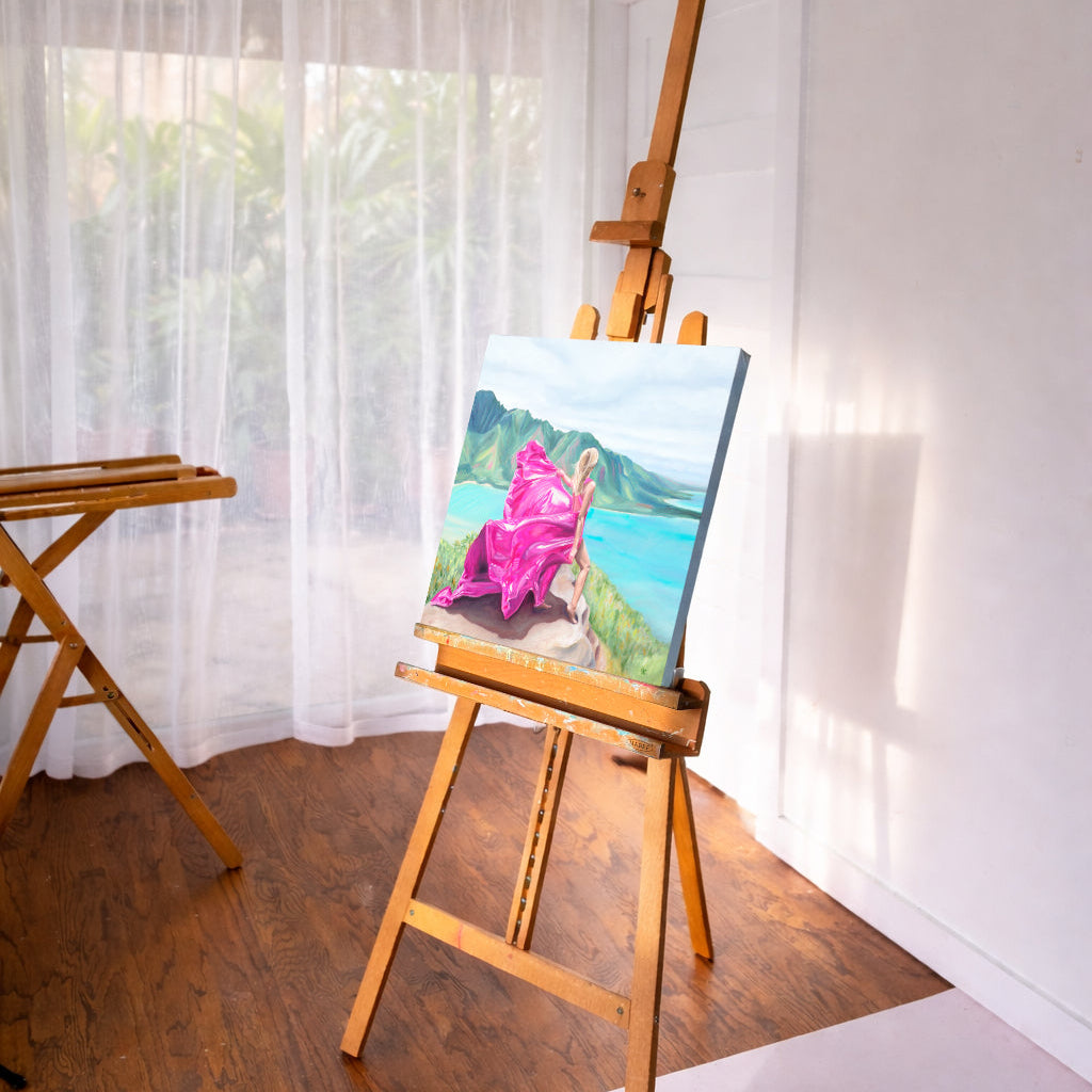 A painting of a woman in a pink dress standing on a mountain in Hawaii overlooking the ocean, on an easel by a window with white curtains.